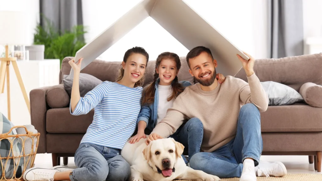 a family of four and a dog seated under a cardboard mock home roof smiling at the camera
